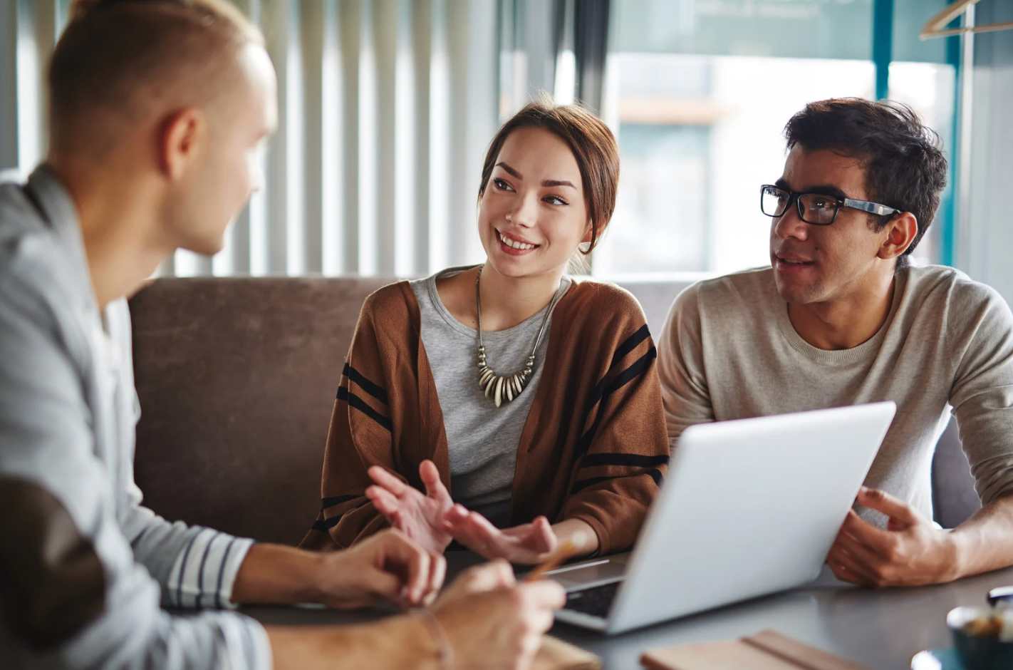 Gegenüber einem Mann sitzen zwei Personen, die ihm interessiert zuhören, am Tisch. Auf dem Tisch sind Dokumente und ein Laptop.